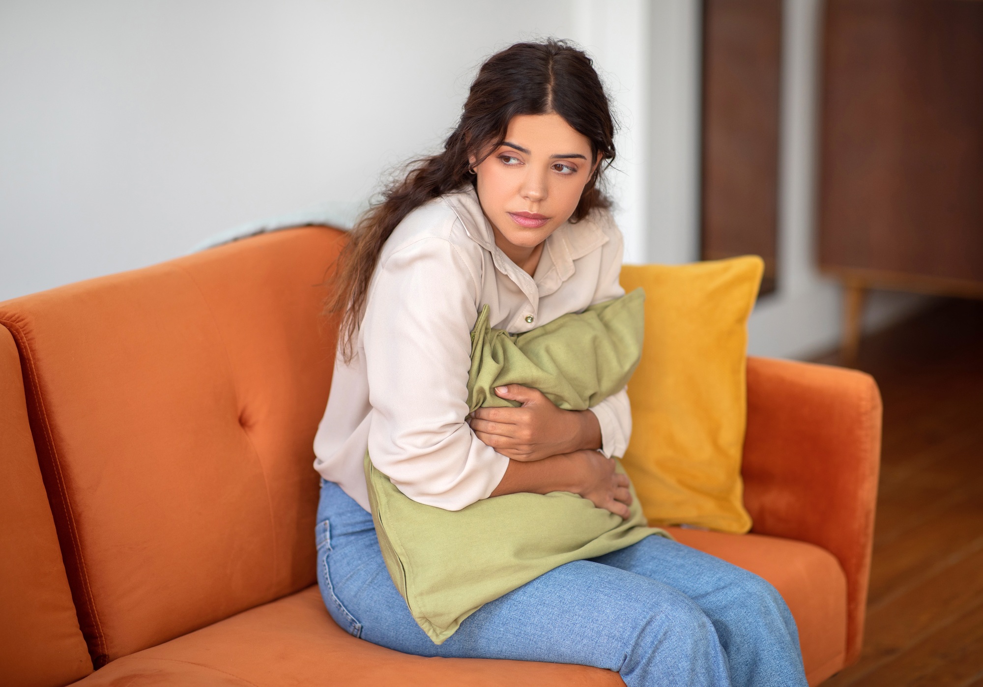 Anxious young woman sitting on sofa, hugging pillow for comfort